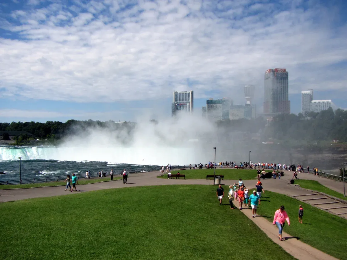 Visitors walking the riverside path near Goat Island with Horseshoe Falls mist behind them on a Niagara Falls tour from NYC, New York